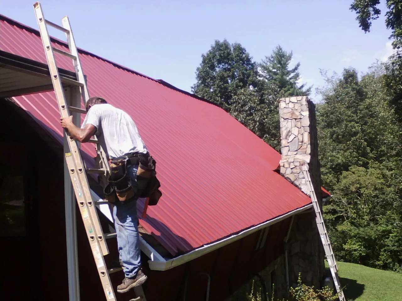 Roofer on a ladder beside a red metal roof and stone chimney — Soffit & Fascia Repair by Roofing Brothers in Blessing